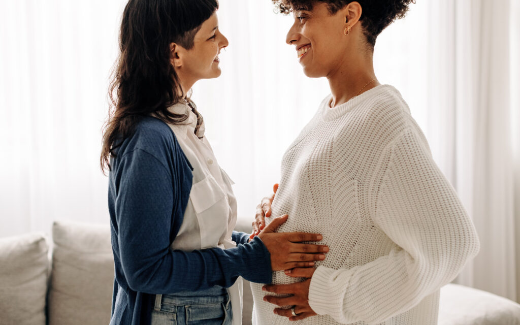 To smilende kvinner vendt mot hverandre, begge holder på kvinnen til høyre sin gravide mage