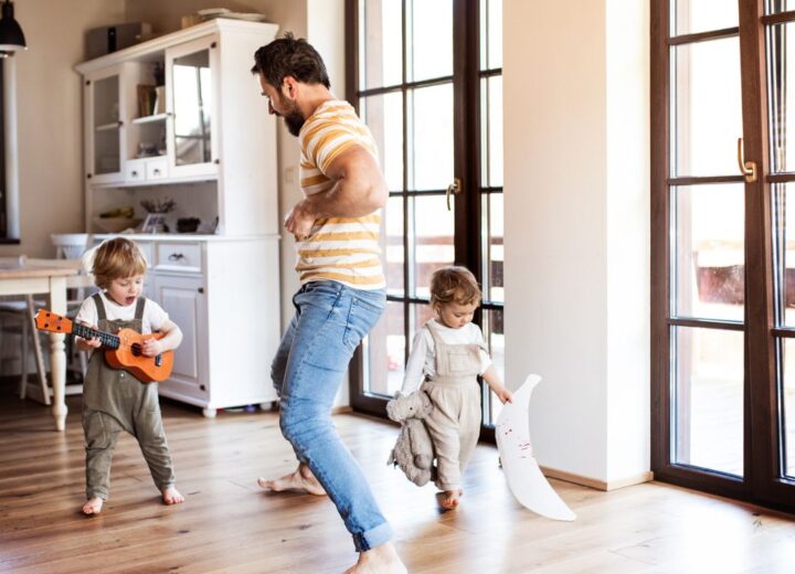 Mann og to barn i en stue. Det ene barnet spiller gitar og synger, det andre barnet bærer på leker. Mannen står i midten og danser.