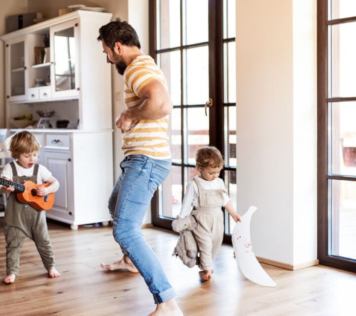 Mann og to barn i en stue. Det ene barnet spiller gitar og synger, det andre barnet bærer på leker. Mannen står i midten og danser.