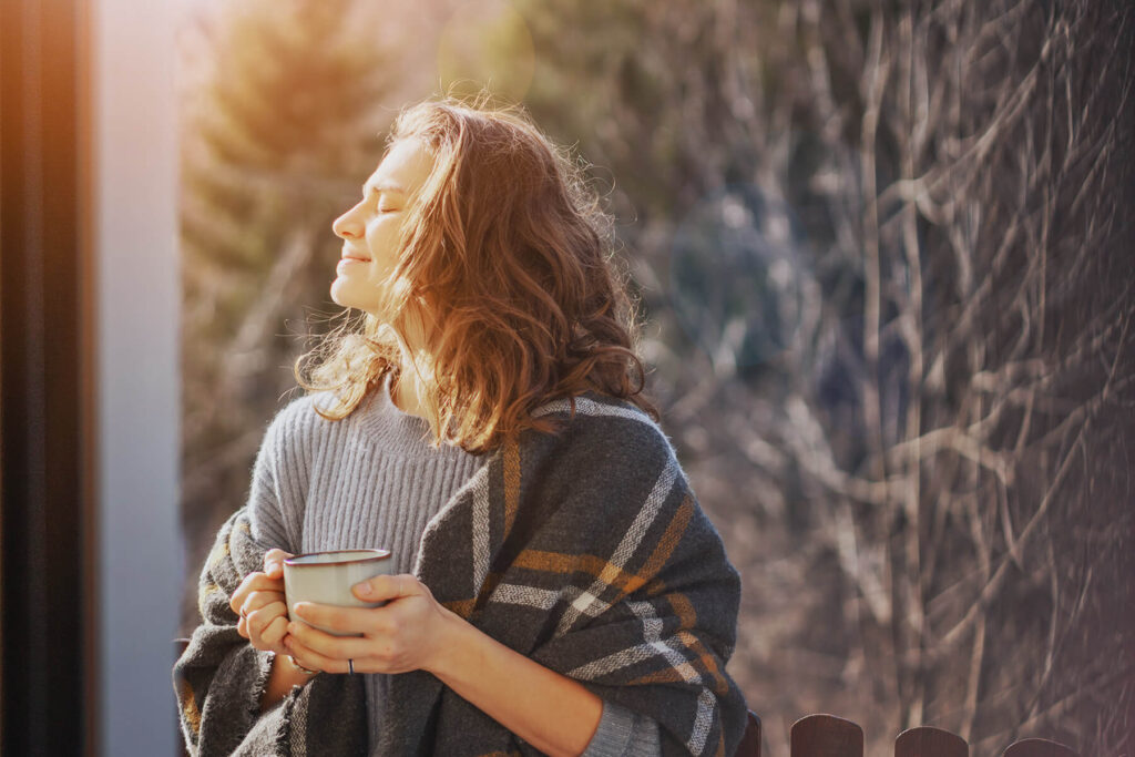Kvinne står med pledd og kaffekopp og nyter solen i ansiktet