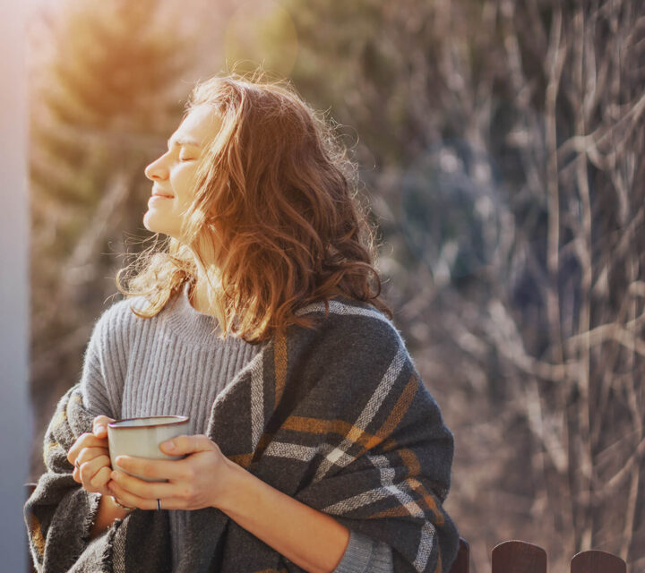 Kvinne står med pledd og kaffekopp og nyter solen i ansiktet